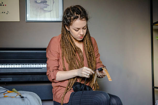 In a cozy apartment corner, a young woman with natural dreadlocks carefully assembles a handmade bow from bamboo, string, and black rope. The image captures a moment of focused craftsmanship and self-reliance.