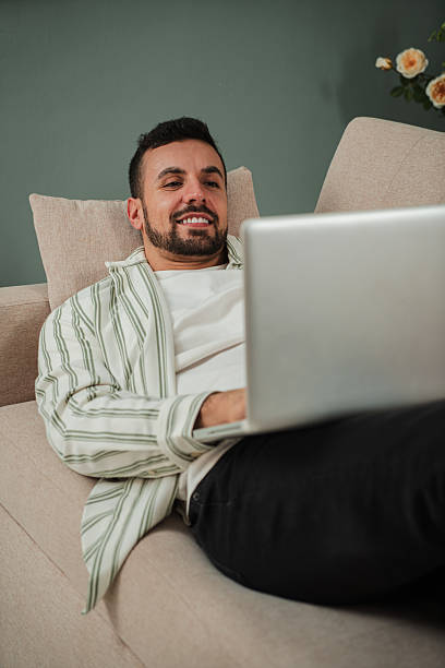 istock Vertical. Handsome Young Adult Man Using Laptop While Sitting On Couch At Home. Smiley Guy Working Remotely On Computer Laying On A Sofa At Cozy Apartment Living Room. Happy Male Enjoying Online Job. 2210274928