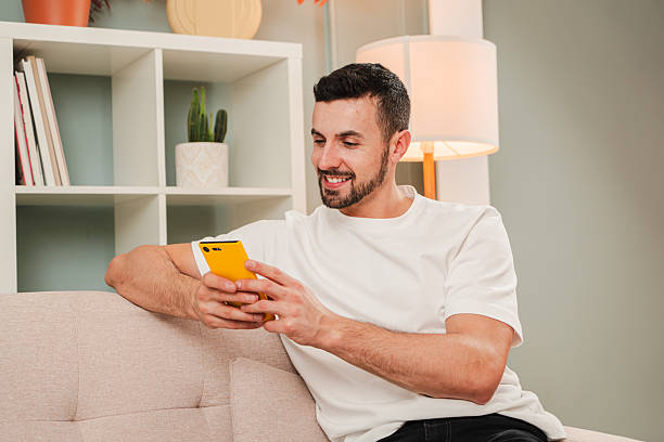 istock Young Man Sitting Comfortably on Couch Using Smartphone with a Bright Yellow Case While Smiling and Engaged in Content. Guy Relaxing in Cozy Living Room with the Social Media App and a Mobile Phone 2210275956