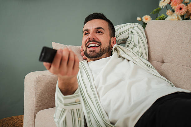 istock Happy man relaxing on a sofa while watching television, enjoying his free time at home. Perfect moment for leisure, entertainment, and personal time with a smile on his face and remote in hand. 2210276008