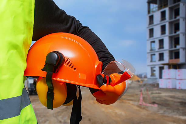 istock Construction worker in a high-visibility vest holding an orange safety helmet and protective gear at a building site. 2210276575