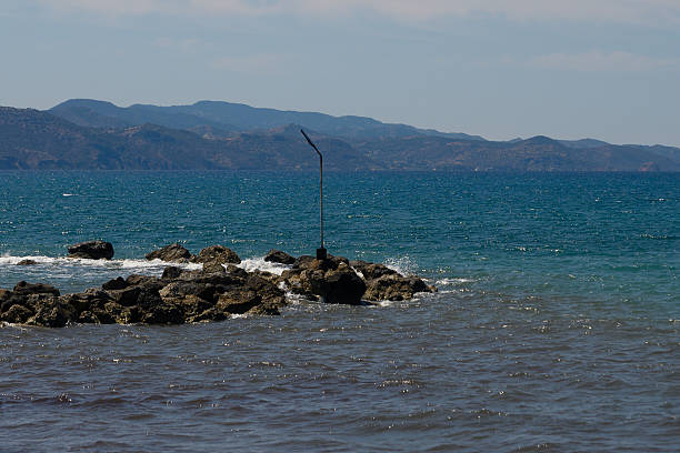 Gentle waves lap against a rocky shoreline, as a solitary pole stands sentinel by the water's edge. The distant mountains are bathed in soft sunlight, enhancing the tranquil setting.