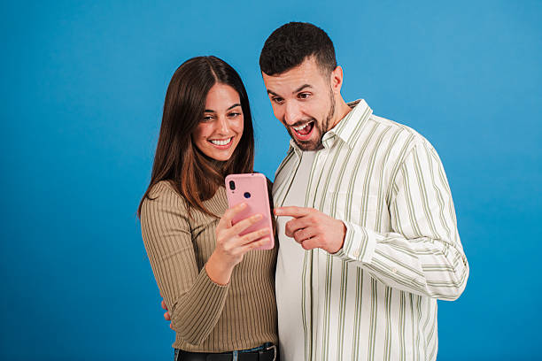 istock Caucasian couple having fun using a mobile phone to browse on internet with an application at blue studio background. Two friends betting online with a cellphone app on a turquoise colour backdrop. 2210279130