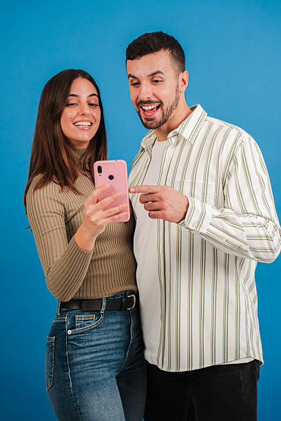 istock Vertical. Caucasian couple having fun using a cell phone to browse on internet with an app at blue studio background. Two friends betting online with a cellphone app on a turquoise colour backdrop. 2210279259