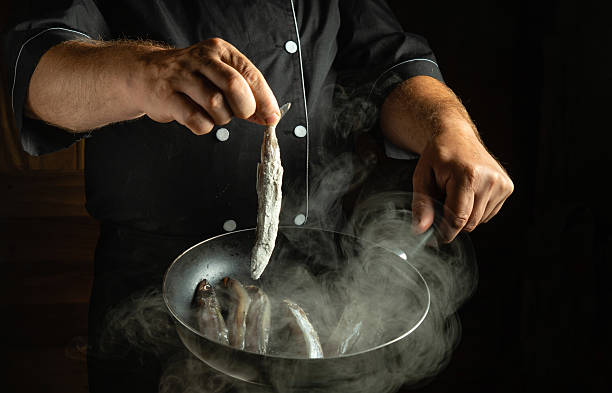istock A chef skillfully holds a fish above a pan in a dark kitchen, while steam rises from the sizzling food. The atmosphere is filled with the aroma of freshly cooked seafood 2210289377
