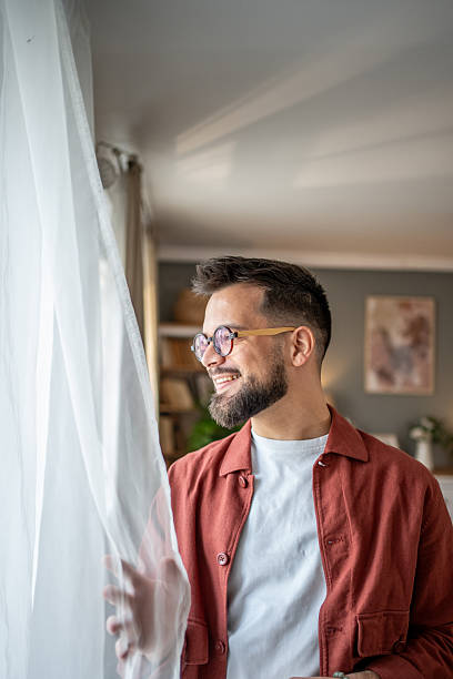 istock Smiling bearded man with glasses looking out the window at home 2210289788