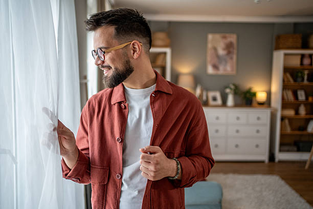 istock Smiling young man opening curtains and looking out the window at home 2210289859