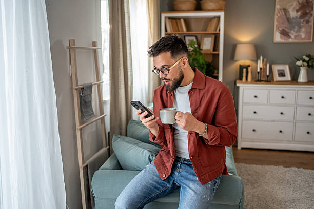 istock Young man sitting on sofa reading news on smartphone and drinking coffee 2210289936