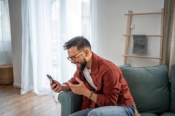 istock Happy man sitting on sofa using smartphone, browsing internet and social media 2210290484