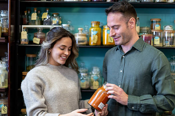 Young smiling shopkeeper showing jar of spices to female customer in zero waste store, small business and healthy lifestyle concept