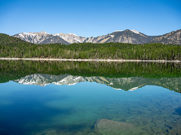 A calm alpine lake in Germany perfectly mirrors the forest and rugged snow-capped mountains under a clear blue sky. The peaceful scenery reflects natural beauty and untouched wilderness.