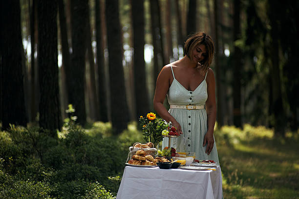A woman arranges a picnic table with fresh food and flowers surrounded by lush forest scenery.