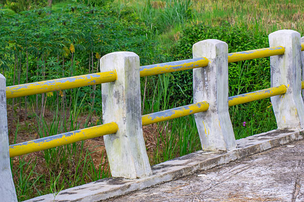 Concrete fence with yellow metal rails, slightly weathered, stretches alongside a pathway. The background features lush green grass and bushes, creating a vibrant natural setting. The combination of weathered paint and greenery suggests an outdoor, rural environment.