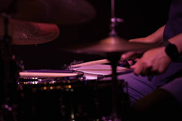 A close-up of a drummer's hands holding drumsticks mid-performance. The dark background and focused lighting create a dynamic atmosphere, highlighting the motion of the drumming.