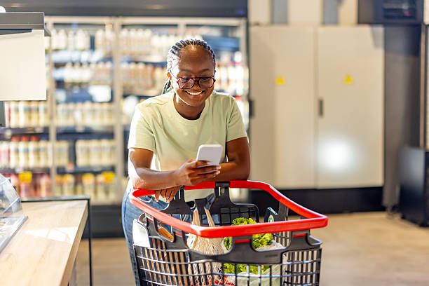 istock Smiling Shopper Using Smartphone While Grocery Shopping with Fresh Produce in Cart 2210300197