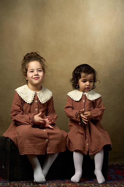 istock Two young sisters in matching brown dresses with lace collars sit on black blocks 2210310571