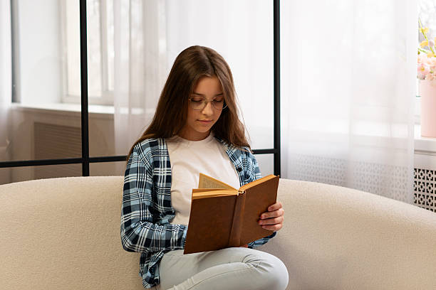 istock Girl wearing glasses sitting on sofa reading book calmly. 2210332115