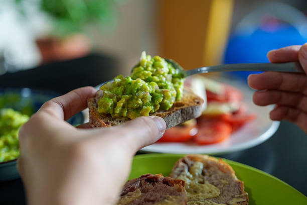 A close-up view of hands spreading mashed avocado onto toasted sourdough bread with melted cheese, featuring a blurred background of tomato salad.