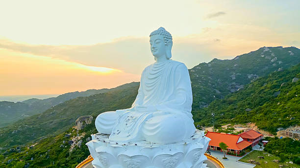 A grand white Buddha statue sits peacefully atop a decorative lotus pedestal, surrounded by lush green hills and bathed in the golden hues of a tranquil sunset sky, near Linh Phong Pagoda.