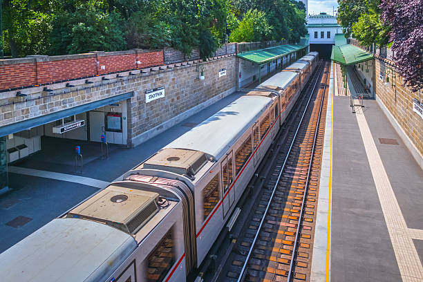 A Vienna metro train arrives at Stadtpark station, an open-air underground station surrounded by greenery and historic stone walls. The platform is empty, and the train's modern design contrasts with the classic architecture of the station.