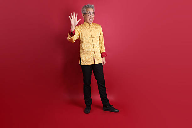 Happy Chinese new year. Asian Chinese senior man wearing golden traditional cheongsam qipao or changshan dress with gesture of Counting number or calculation isolated on red background.