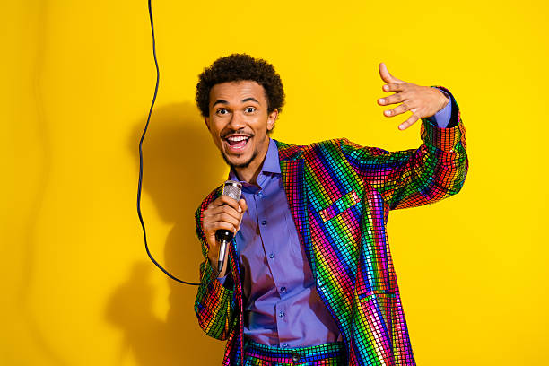 istock Young man in colorful disco suit singing with microphone in vibrant yellow background, expressing fun and celebration at a joyful party 2210342356