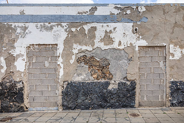 This image captures a weathered wall in Arrecife, Lanzarote, featuring three sealed doorways. The surfaces show layers of peeling paint, exposed brickwork, and plaster repairs, reflecting the passage of time and structural modifications. The lower section of the wall consists of dark, textured stone, contrasting with the lighter plaster above. The paved sidewalk in the foreground adds context to the urban setting, highlighting the evolving architectural landscape of the Canary Islands.
