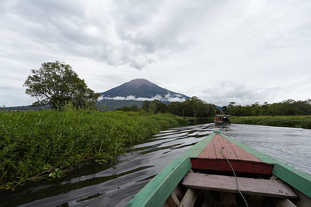 A scenic view from the bow of a wooden boat navigating a calm river or canal surrounded by lush green wetlands, with Mount Kerinci — the highest volcano in Indonesia — standing majestically in the background. The landscape is covered in clouds, adding a dramatic and tranquil atmosphere. This image is perfect for illustrating nature tourism, ecotourism, volcano landscapes, and traditional travel in Sumatra.