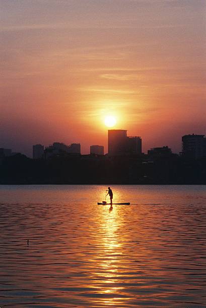 A person is paddleboarding on West Lake during sunset in Hanoi, Vietnam. The city skyline is visible in the background, reflecting on the calm water. For editorial use only.
