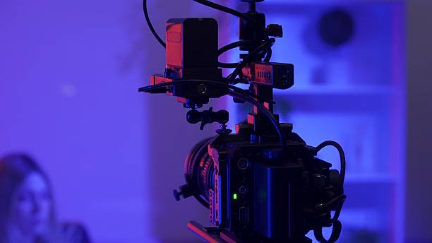 istock Camera on a tripod against a blurred background of a female presenter in a dark studio in pink, blue neon lights. 2210358528