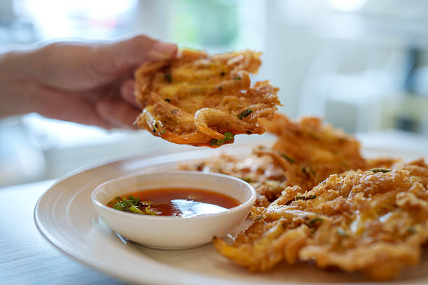 Close-up of a hand picking up a golden crispy prawn fritter served with a spicy dipping sauce. A popular street food snack in Penang, Malaysia.