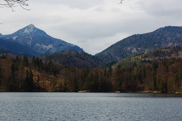The somber, still surface of Hechtsee lies before a dense, dark forest, all watched over by imposing, cloud-covered mountain peaks in the Austrian Alps