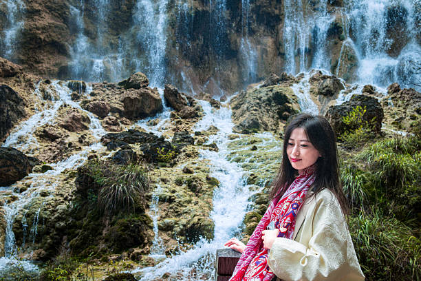 A woman stands in front of a waterfall, dressed in a white outfit with a red scarf, showing a bright smile against the stunning natural backdrop
