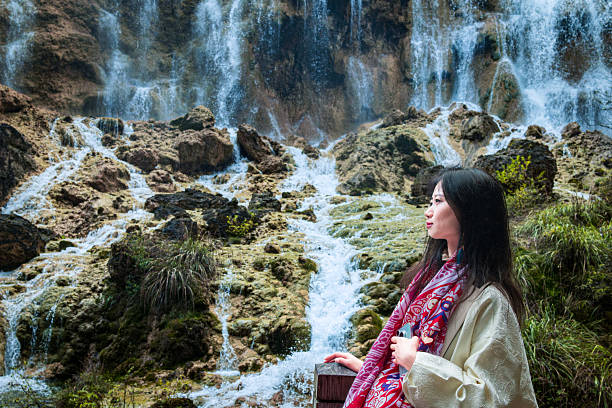 A woman stands in front of a waterfall, dressed in a white outfit with a red scarf, showing a bright smile against the stunning natural backdrop