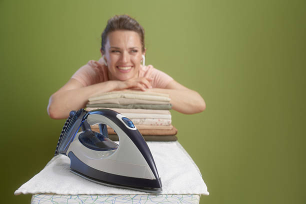 A smiling woman rests on folded clothes with an iron in the foreground, set against a muted olive green background. She appears relaxed and happy.