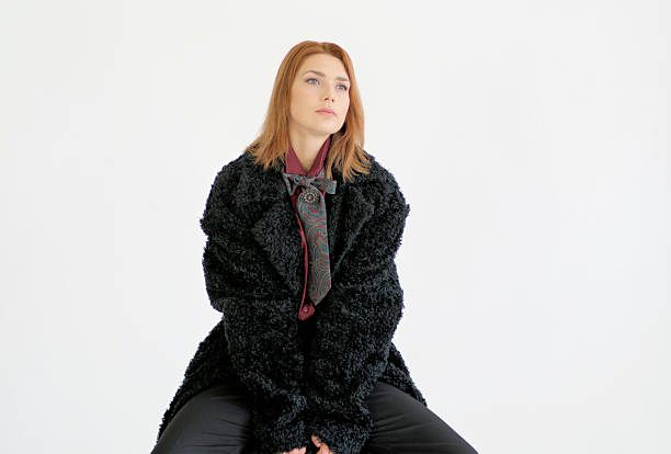 Studio portrait of a young adult Caucasian woman wearing a black textured winter coat, seated confidently against a clean white backdrop in a minimalist setting.