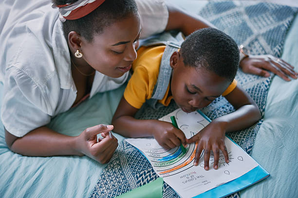 Mother, boy and coloring book on bed with learning, development and support with bonding in home. African woman, child and paper in bedroom with drawing, art and relax with education in family house