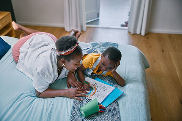 istock Black woman, child and coloring book on bed with learning, development and support with bonding in home. Mother, boy and paper in bedroom with drawing, art and relax with education in family house 2210374850