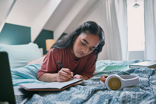istock University student, writing and study with notebook on bed for exam revision, planning essay and thesis research. Laptop, girl and learning college information in home for scholarship and assessment 2210374915