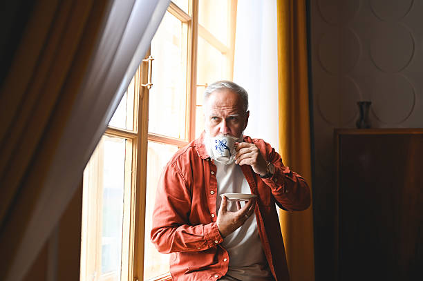 istock Senior man with white beard drinking tea and looking out the window while standing in his home 2210381461