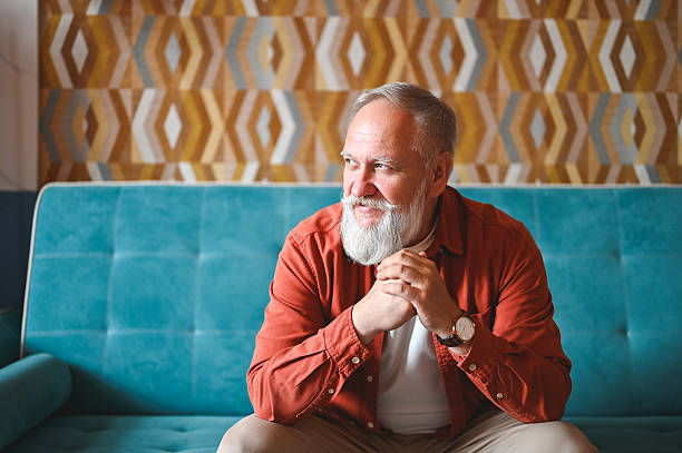 istock Senior man smiling and looking away while sitting on sofa at home 2210381530