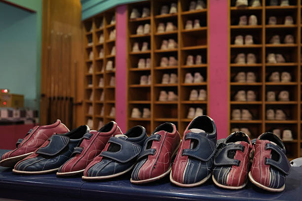 Bowling shoes are lined up on a counter, ready for bowlers to use, with a shoe rack full of more shoes in the background