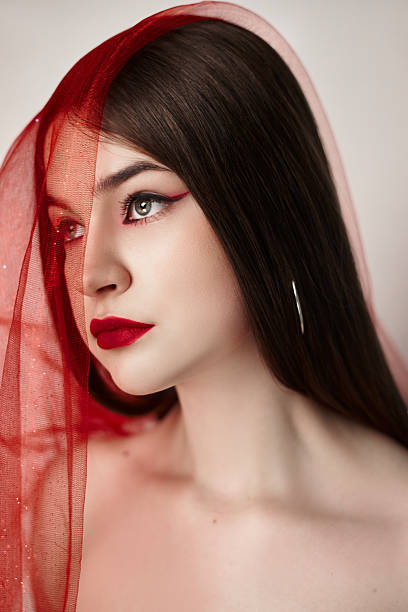 istock woman poses elegantly with a sheer red veil draped over her head, emphasizing her delicate features and bold makeup. soft background enhances her captivating gaze and aesthetic 2210391025
