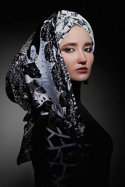 young woman poses confidently with a black and white patterned headscarf draped over her shoulder. studio backdrop is dark, highlighting her features and the intricate design of her scarf.