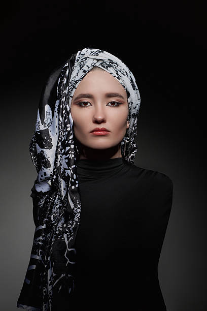 istock woman showcases a bold expression while wearing a patterned black and white headscarf. emphasis is on her features illuminated softly against the dark backdrop, creating a dramatic effect 2210391052