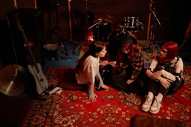 istock Girls Band of Three Young Artists Using Phone while Sitting on Carpet in Dim Studio 2210397813