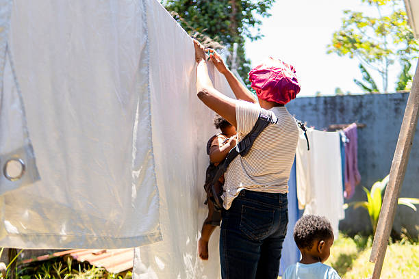 Mother hanging laundry outside while carrying her baby in a carrier with her son watching nearby