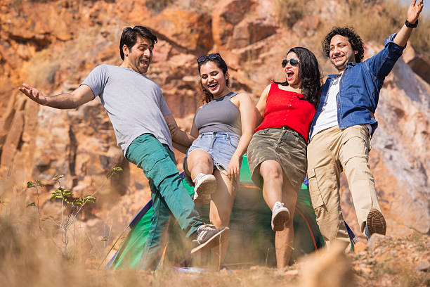 Joyful young couples laughing and playfully standing on one leg while posing outside tent against rocky mountain backdrop during the weekend