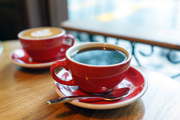 A close-up view of two steaming cups of coffee, one black and one latte with latte art, served in matching red cups on a wooden café table.