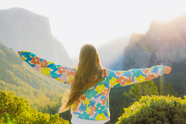 Rear view of happy serene female with long hair in colourful fleece shirt raising arms at the scenic viewpoint of summer sunrise above majestic mountain range with forest and waterfall in Yosemite National park, California, the United States